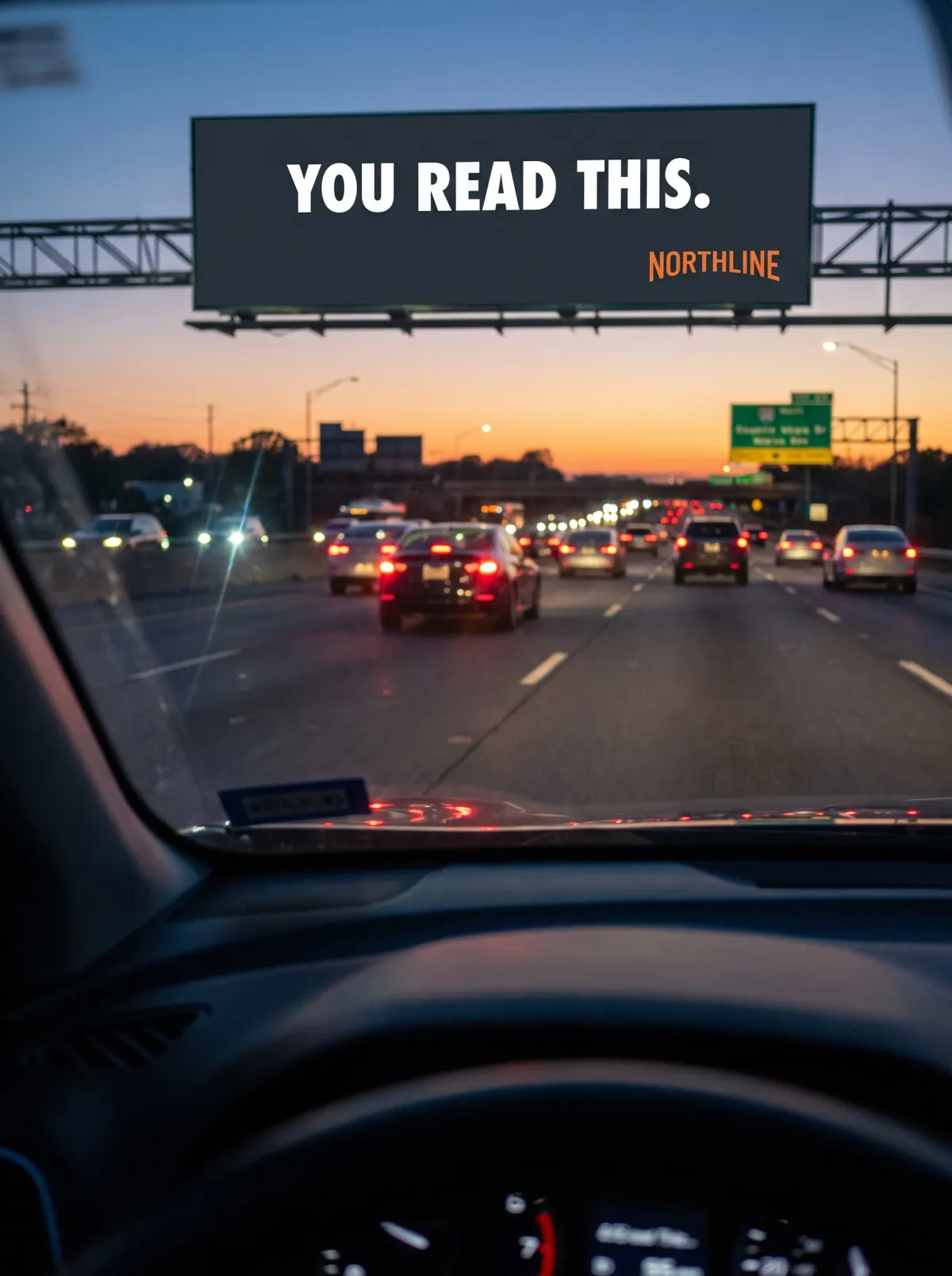 A driver on a highway at dusk reading a Northline billboard through the windshield
